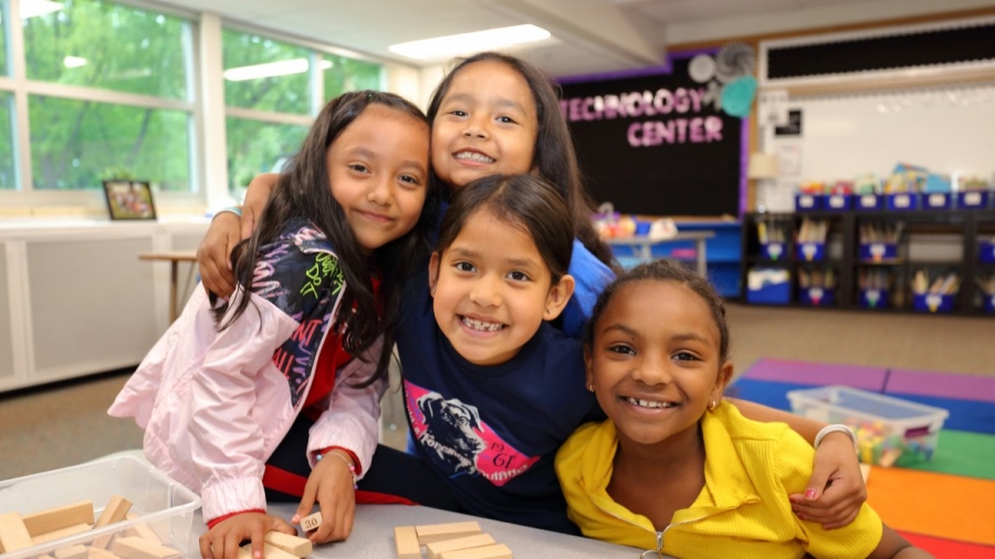 Image of 4 young kids hugging, in a classroom, huddled around a table playing blocks. They look like they're all friends.