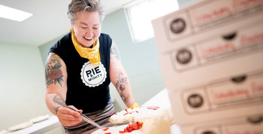 Image of a person (Rachel, one of the owners of Pie & Mighty) using a tweezers to place strawberries as garnish on a pie. There's also a stack of pie boxes in the foreground. 