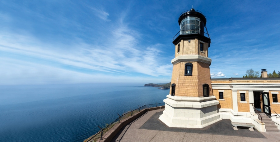 Image of Split Rock Lighthouse, up close. It appears to be up on the top of a hill, overlooking water and the sky, and there's a couple story tall lighthouse that is 20 feet away.