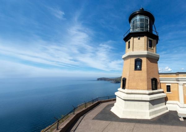 Image of Split Rock Lighthouse, up close. It appears to be up on the top of a hill, overlooking water and the sky, and there's a couple story tall lighthouse that is 20 feet away.