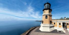 Image of Split Rock Lighthouse, up close. It appears to be up on the top of a hill, overlooking water and the sky, and there's a couple story tall lighthouse that is 20 feet away.