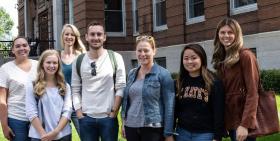 A group of college students, mostly women, standing in front of a brick college building, smiling. 