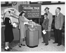 A woman inserts a paper bill into a large glass globe to support the Spirit of 1776 Freedom Train, as two men, a boy, and another woman look on.  Circa 1948.